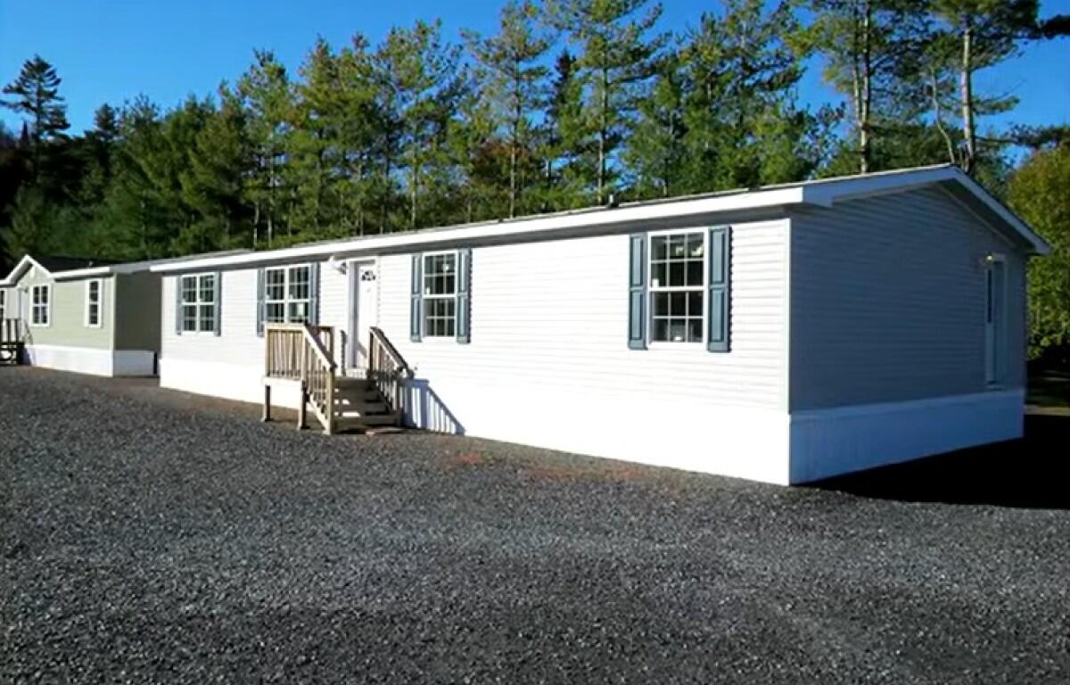 White manufactured home with blue shutters on a gravel lot, trees in the background, and a small wooden staircase at the front door.