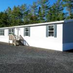 White manufactured home with blue shutters on a gravel lot, trees in the background, and a small wooden staircase at the front door.