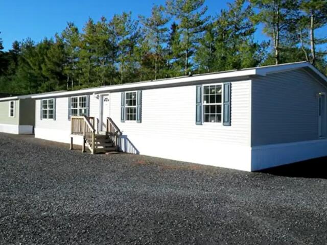 White manufactured home with blue shutters on a gravel lot, trees in the background, and a small wooden staircase at the front door.