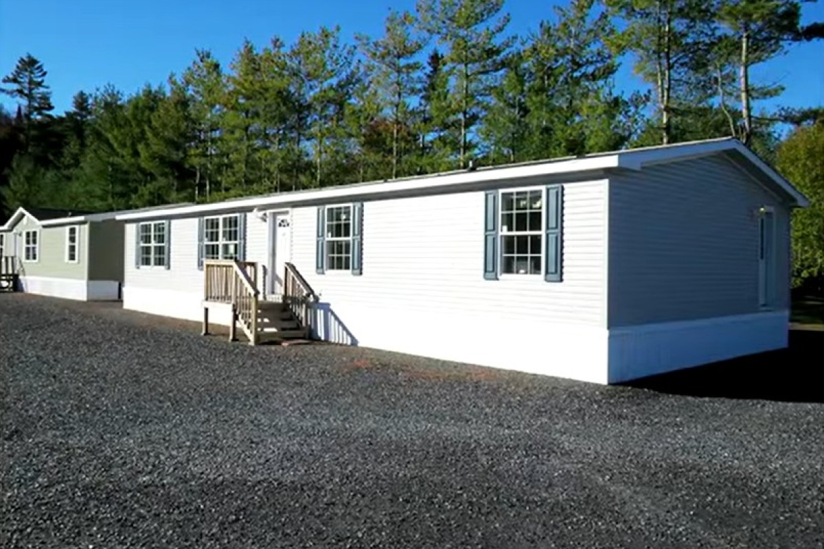 White manufactured home with blue shutters on a gravel lot, trees in the background, and a small wooden staircase at the front door.