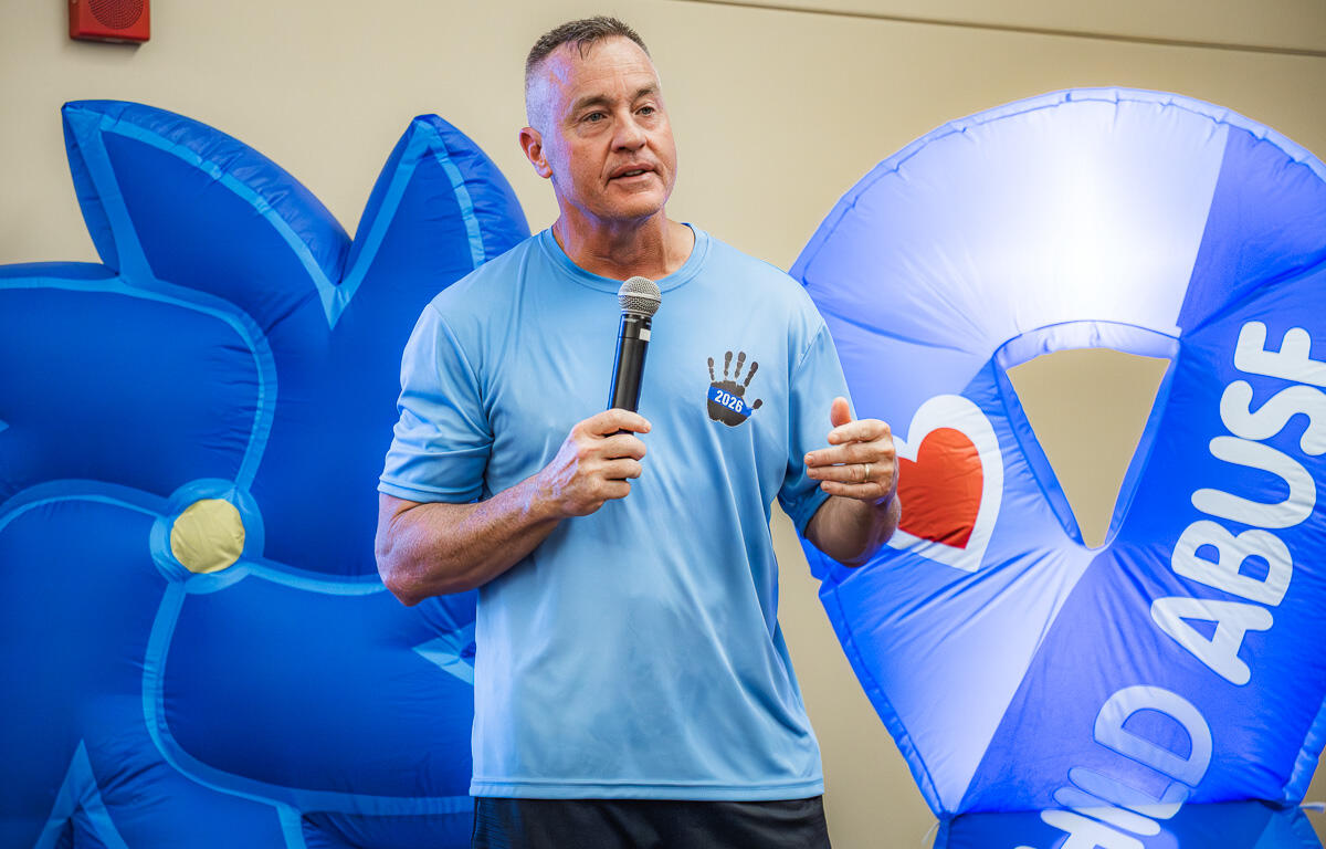 Man speaking into a microphone at an indoor event, wearing a light blue shirt, with large blue inflatables in the background featuring a heart and text.