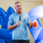 Man speaking into a microphone at an indoor event, wearing a light blue shirt, with large blue inflatables in the background featuring a heart and text.