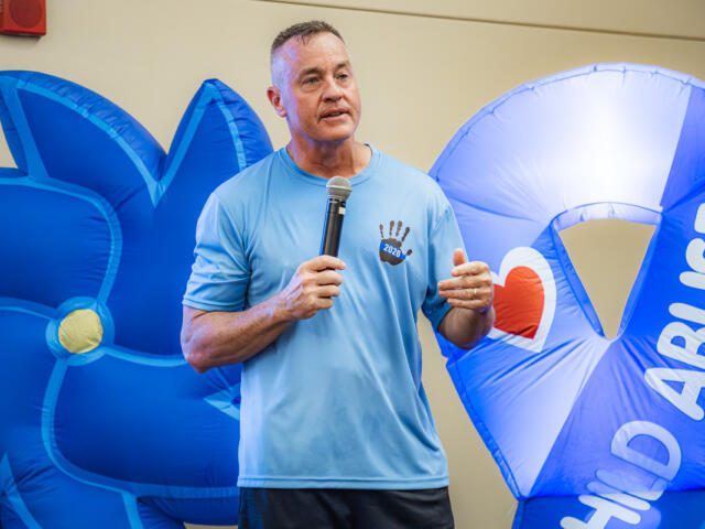 Man speaking into a microphone at an indoor event, wearing a light blue shirt, with large blue inflatables in the background featuring a heart and text.