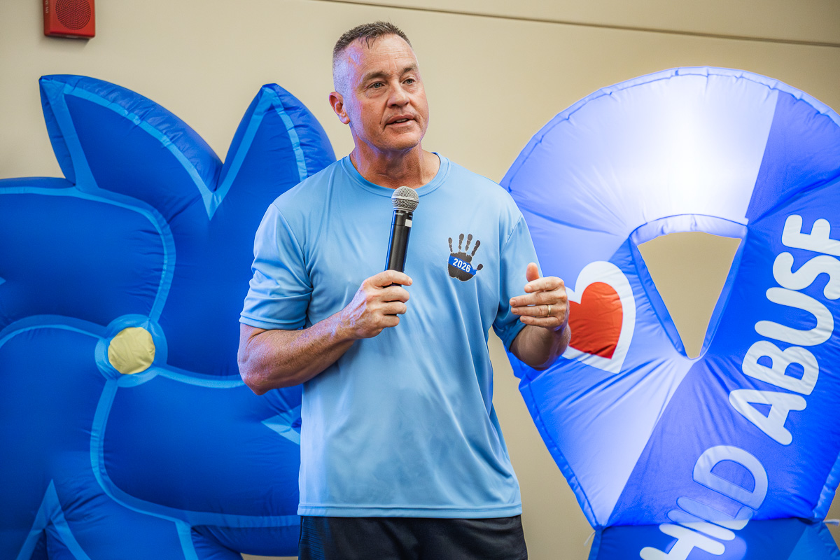 Man speaking into a microphone at an indoor event, wearing a light blue shirt, with large blue inflatables in the background featuring a heart and text.