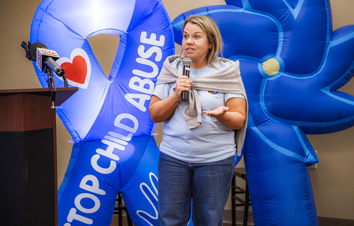 Woman speaks into a microphone at a podium, with large blue inflatable anti-child-abuse display behind her.
