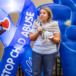 Woman speaks into a microphone at a podium, with large blue inflatable anti-child-abuse display behind her.
