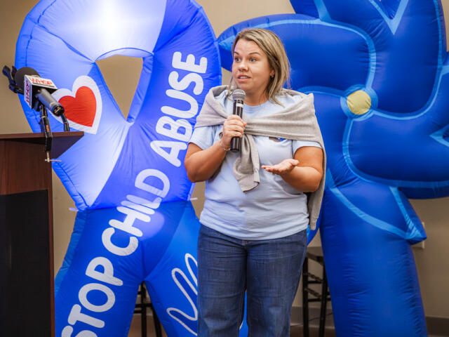 Woman speaks into a microphone at a podium, with large blue inflatable anti-child-abuse display behind her.