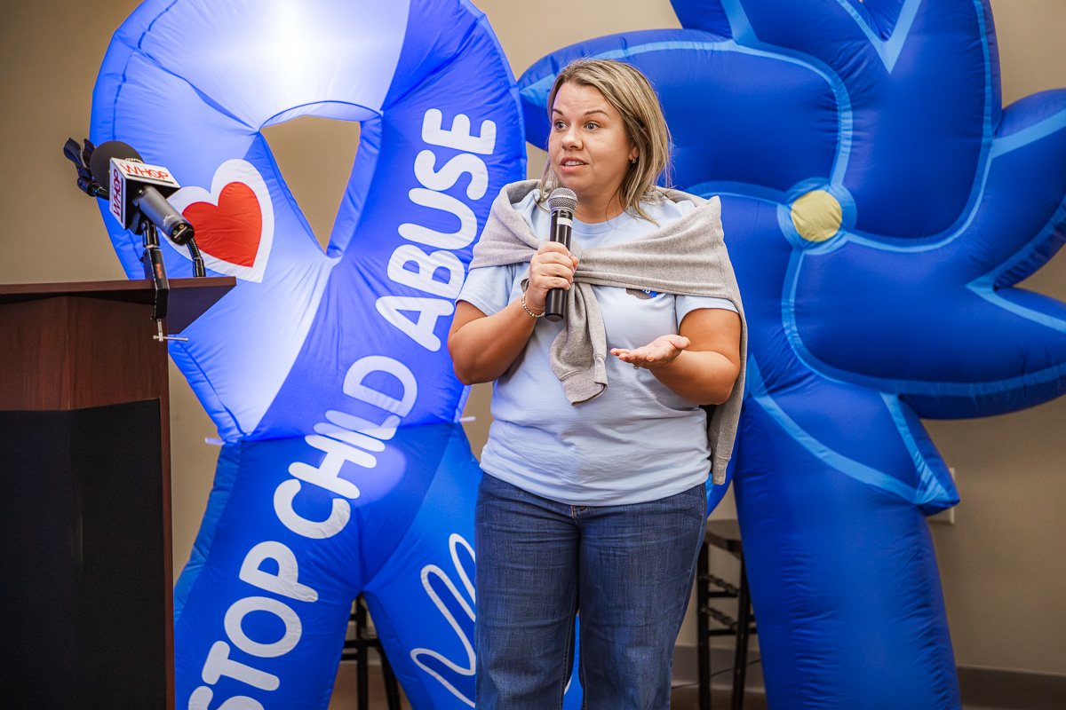 Woman speaks into a microphone at a podium, with large blue inflatable anti-child-abuse display behind her.