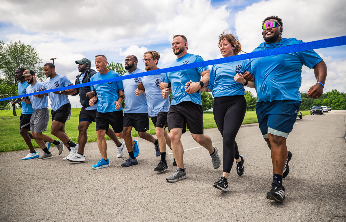 A group of diverse runners in matching blue shirts sprinting together at a race start behind a blue tape line.