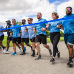 A group of diverse runners in matching blue shirts sprinting together at a race start behind a blue tape line.