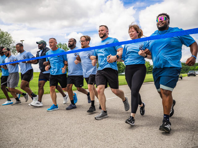 A group of diverse runners in matching blue shirts sprinting together at a race start behind a blue tape line.