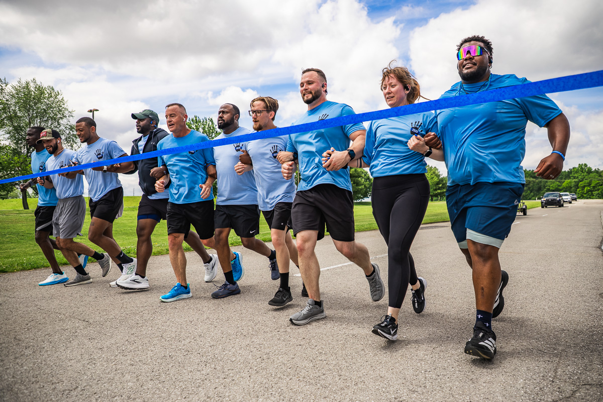 A group of diverse runners in matching blue shirts sprinting together at a race start behind a blue tape line.