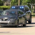 Silver Nissan sedan with its front left door open on a residential street; a dark SUV with blue emergency lights follows behind.