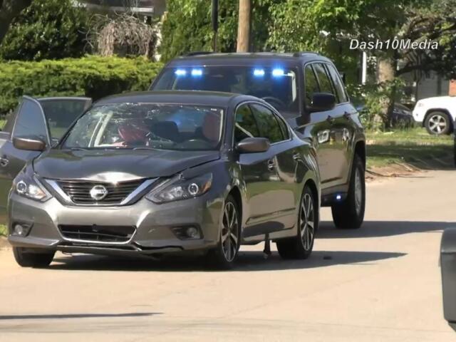 Silver Nissan sedan with its front left door open on a residential street; a dark SUV with blue emergency lights follows behind.