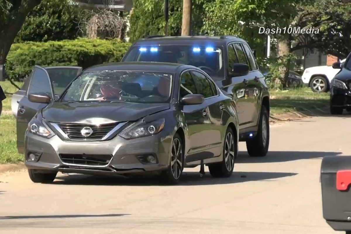 Silver Nissan sedan with its front left door open on a residential street; a dark SUV with blue emergency lights follows behind.