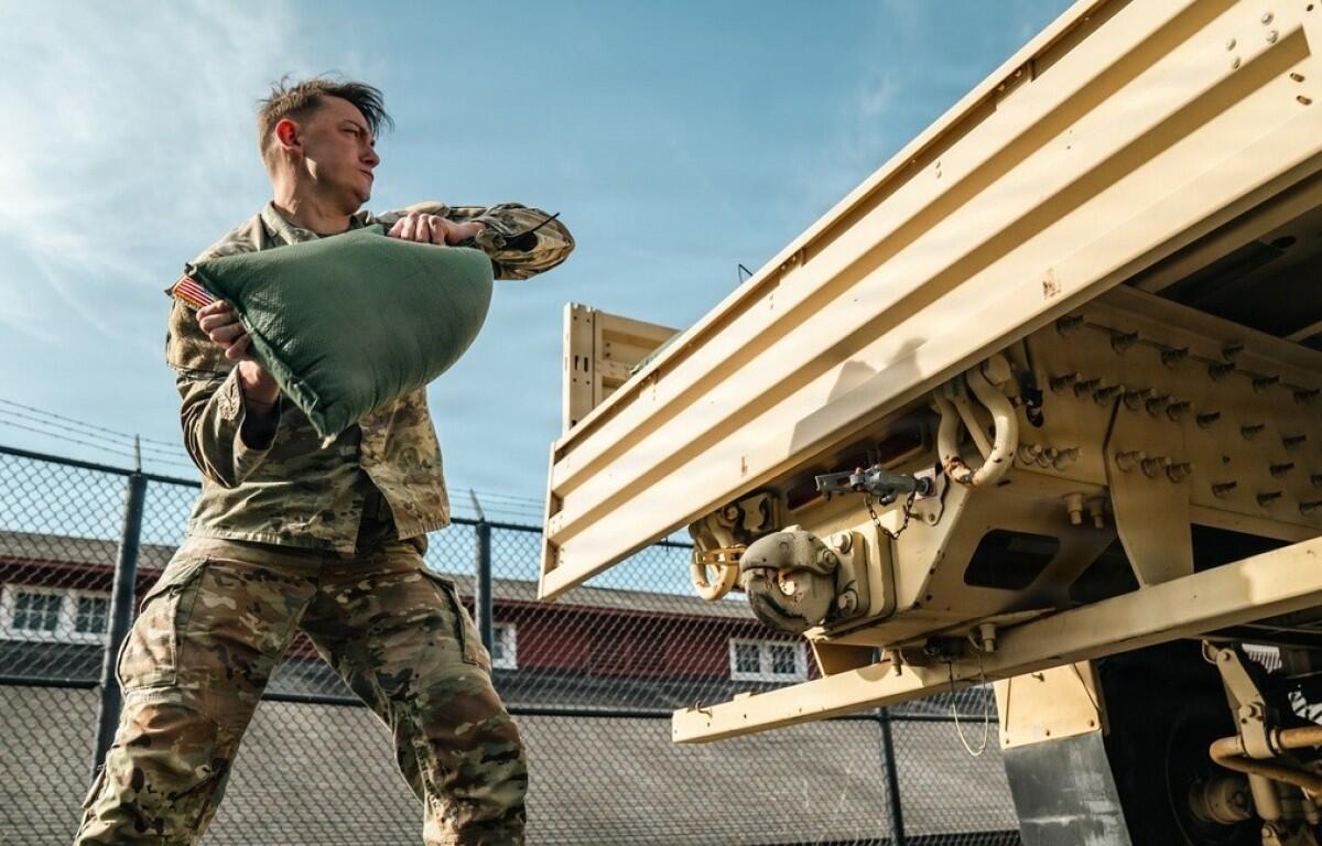 Soldier in camouflage uniforms lifting a green bag toward a large beige military trailer.