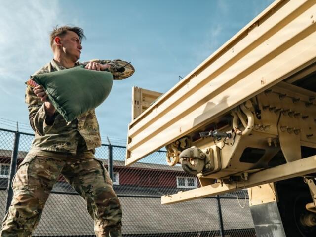 Soldier in camouflage uniforms lifting a green bag toward a large beige military trailer.