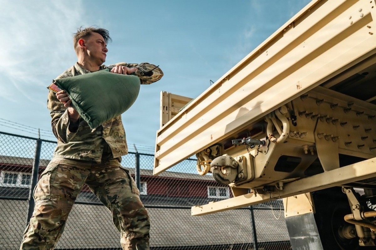 Soldier in camouflage uniforms lifting a green bag toward a large beige military trailer.