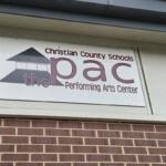 Sign reading 'Christian County Schools PAC' and 'Performing Arts Center' with a building logo on a white plaque above a brick wall