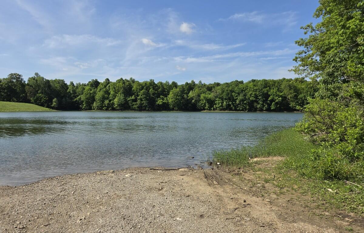 Calm lake with a pebbled shoreline and dense green trees across the water under a blue, partly cloudy sky.