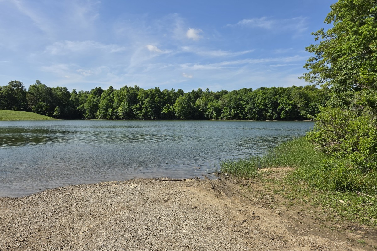 Calm lake with a pebbled shoreline and dense green trees across the water under a blue, partly cloudy sky.