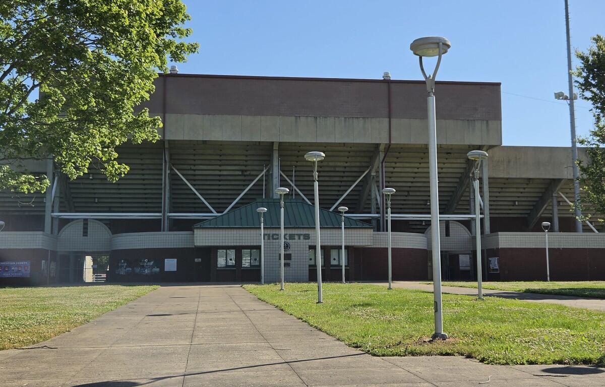 Front view of a large concrete stadium with a green-roofed entrance, a paved walkway leading to the doors, and tall modern street lamps on a grassy plaza.