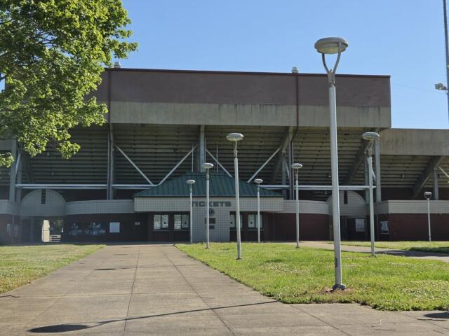 Front view of a large concrete stadium with a green-roofed entrance, a paved walkway leading to the doors, and tall modern street lamps on a grassy plaza.