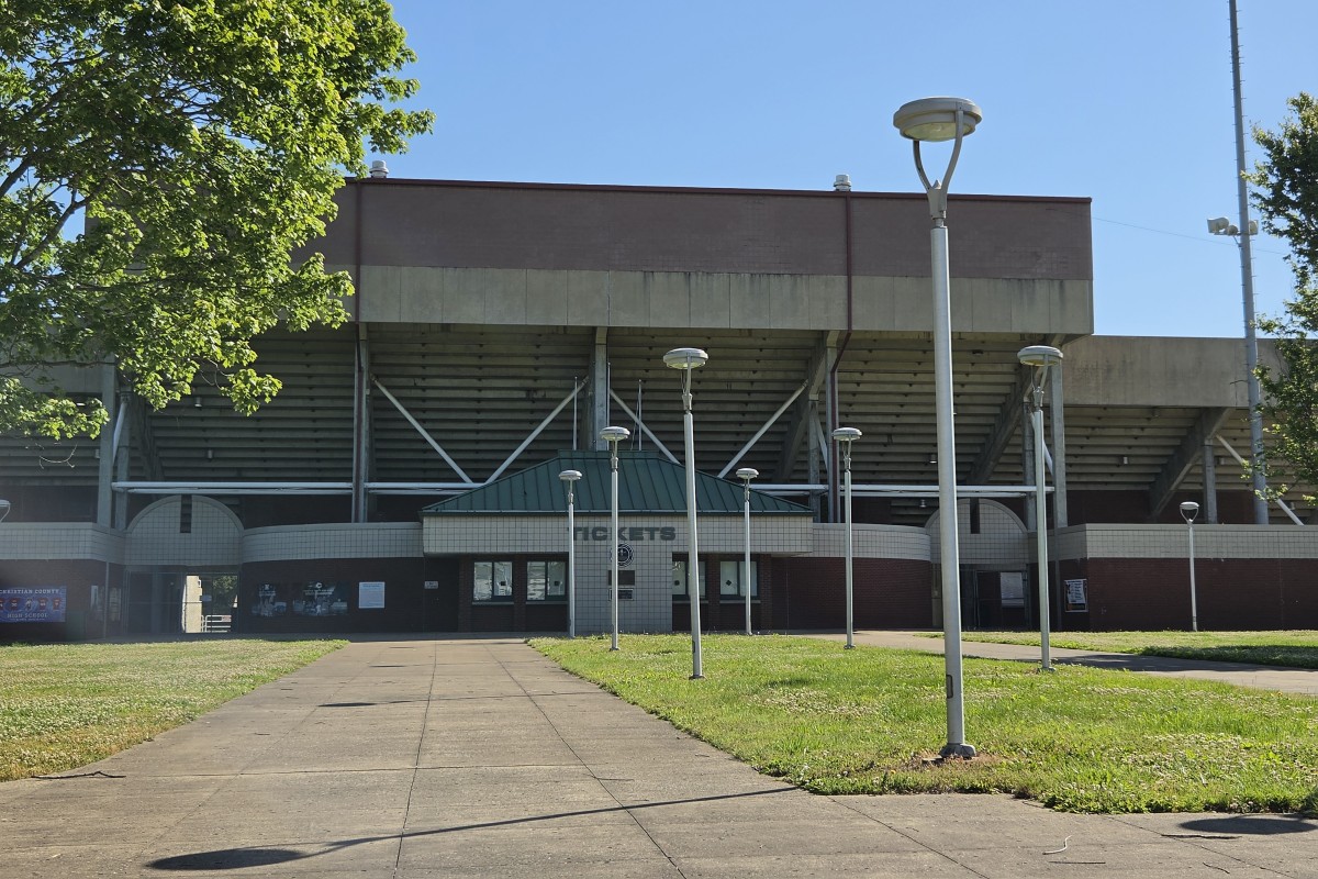 Front view of a large concrete stadium with a green-roofed entrance, a paved walkway leading to the doors, and tall modern street lamps on a grassy plaza.