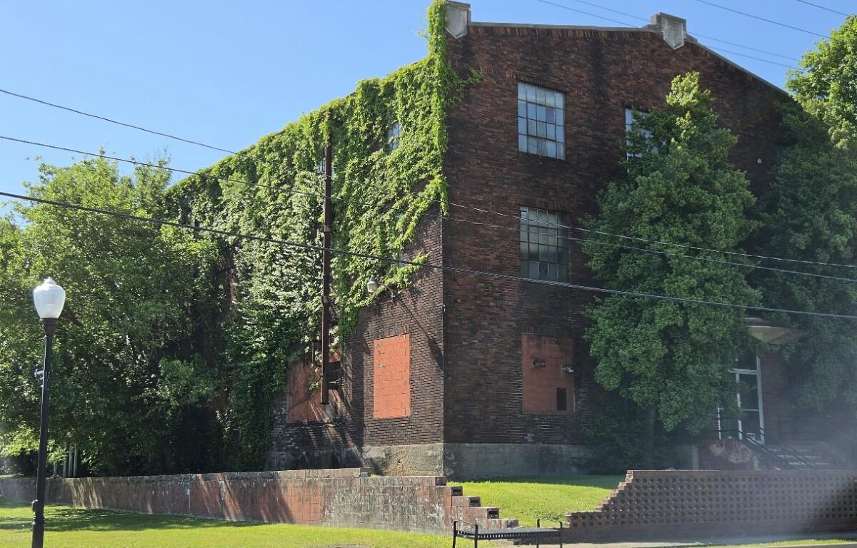 Brick building largely covered in ivy with boarded windows, set in a grassy, park-like area.