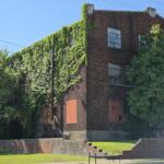 Brick building largely covered in ivy with boarded windows, set in a grassy, park-like area.
