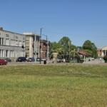 A quiet city street lined with low-rise buildings and parked cars, with a grassy foreground and clear blue sky.