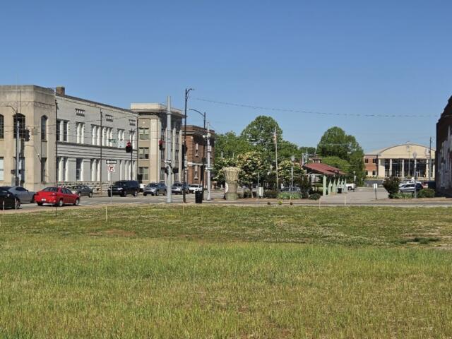 A quiet city street lined with low-rise buildings and parked cars, with a grassy foreground and clear blue sky.