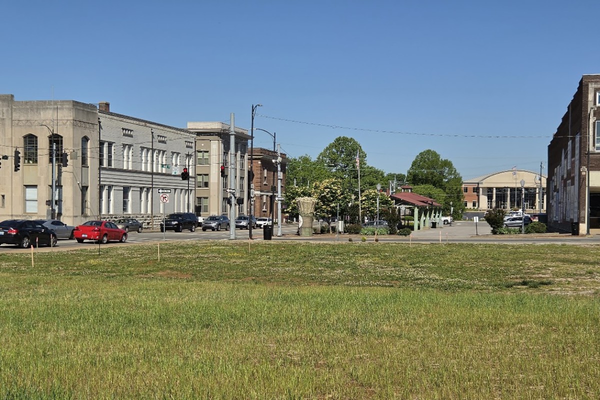 A quiet city street lined with low-rise buildings and parked cars, with a grassy foreground and clear blue sky.