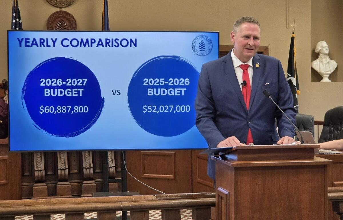 Public official in a navy suit and red tie speaks at a lectern beside a large screen titled 'Yearly Comparison,' showing 2026-2027 budget $60,887,800 vs 2025-2026 budget $52,027,000.