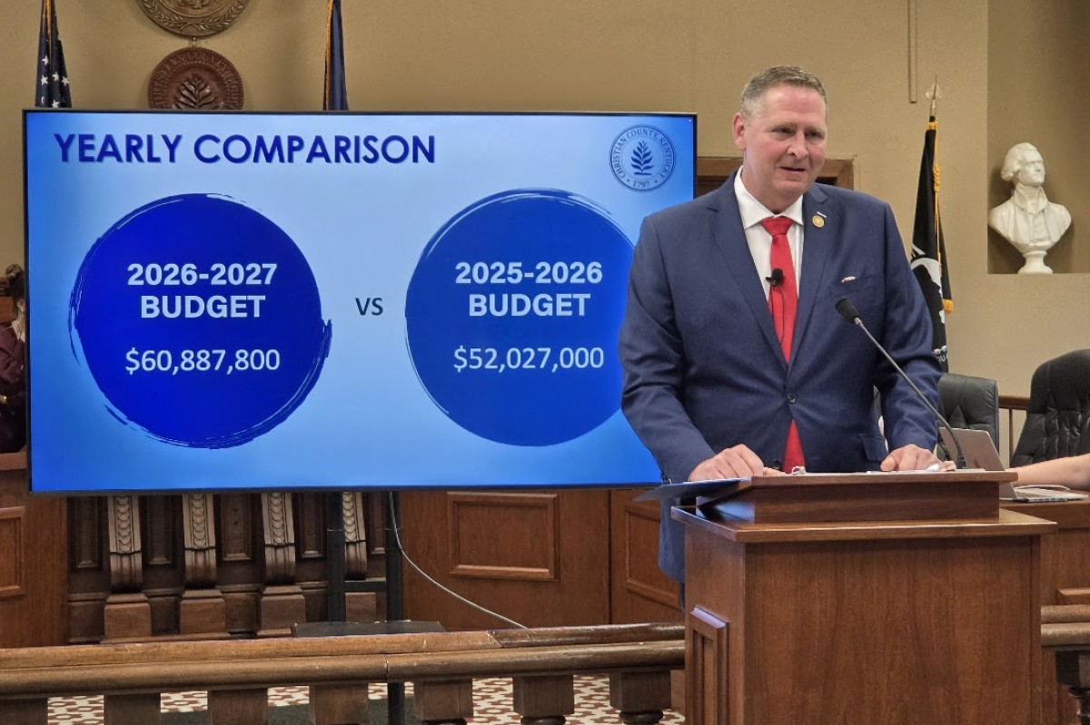Public official in a navy suit and red tie speaks at a lectern beside a large screen titled 'Yearly Comparison,' showing 2026-2027 budget $60,887,800 vs 2025-2026 budget $52,027,000.