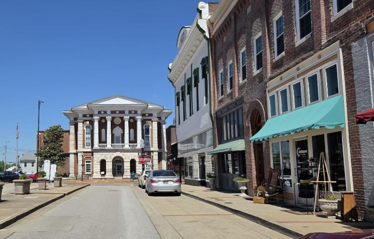 Street view of a small town with a neoclassical courthouse-style building on the left and brick storefronts with green awnings on the right, under a blue sky.
