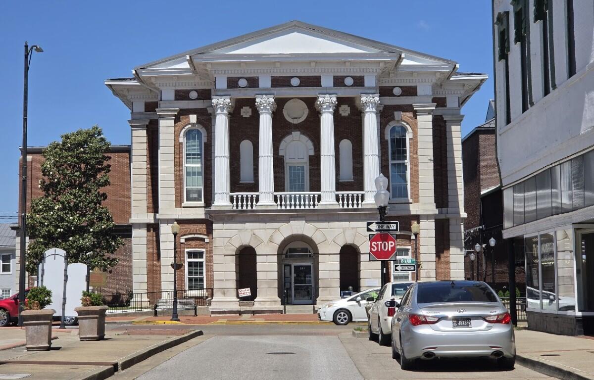 Neoclassical brick courthouse with four white columns and arched entrance on a sunny street, cars parked along the curb.