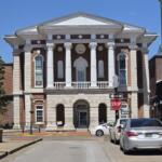 Neoclassical brick courthouse with four white columns and arched entrance on a sunny street, cars parked along the curb.