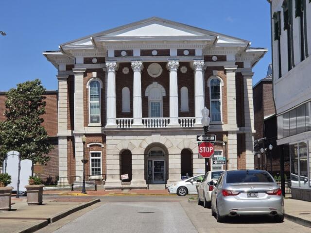 Neoclassical brick courthouse with four white columns and arched entrance on a sunny street, cars parked along the curb.