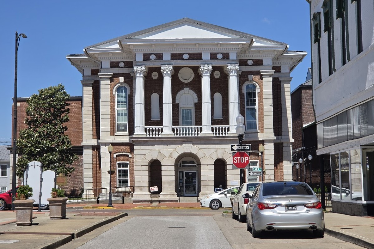 Neoclassical brick courthouse with four white columns and arched entrance on a sunny street, cars parked along the curb.