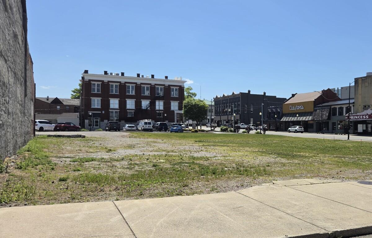 Vacant lot with sparse grass and a sidewalk, urban street and brick building storefronts in the background on a sunny day.