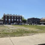 Vacant lot with sparse grass and a sidewalk, urban street and brick building storefronts in the background on a sunny day.