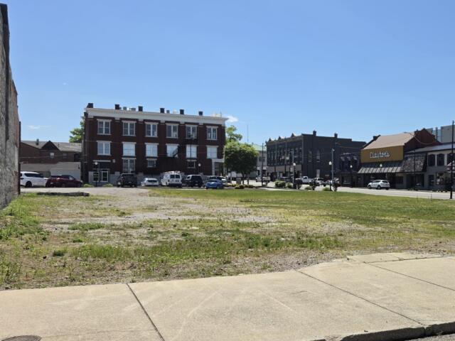 Vacant lot with sparse grass and a sidewalk, urban street and brick building storefronts in the background on a sunny day.
