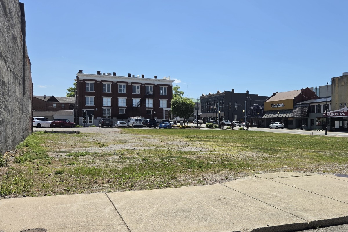 Vacant lot with sparse grass and a sidewalk, urban street and brick building storefronts in the background on a sunny day.