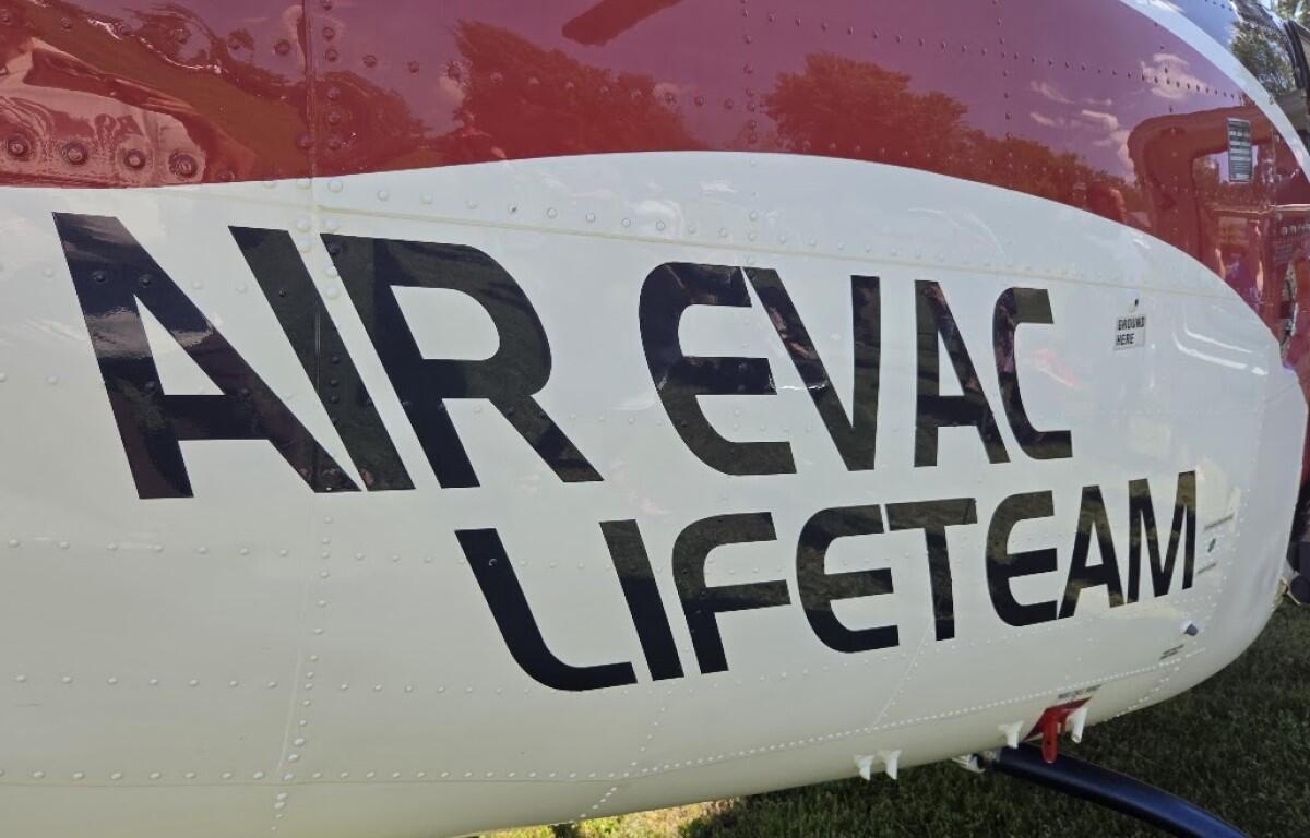 Helicopter fuselage with bold black lettering reading 'AIR EVAC LIFETEAM' on a white and red body, parked on grass.