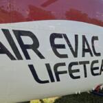 Helicopter fuselage with bold black lettering reading 'AIR EVAC LIFETEAM' on a white and red body, parked on grass.