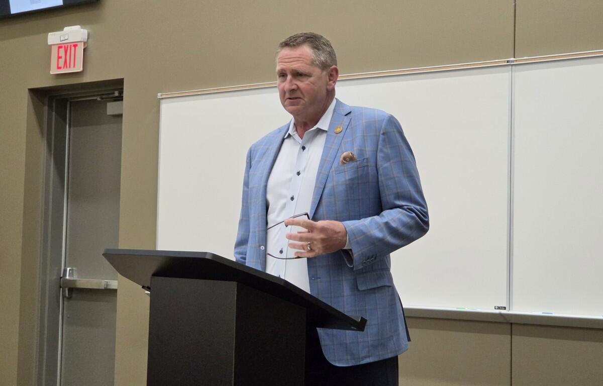 Man in a blue checkered blazer stands at a podium giving a lecture in a classroom setting.