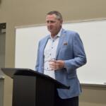 Man in a blue checkered blazer stands at a podium giving a lecture in a classroom setting.
