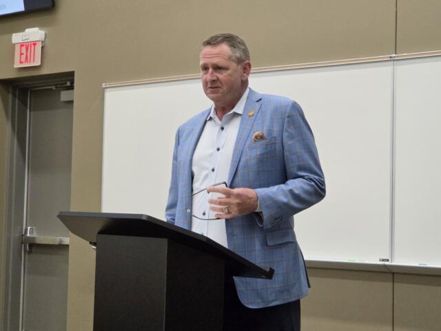 Man in a blue checkered blazer stands at a podium giving a lecture in a classroom setting.