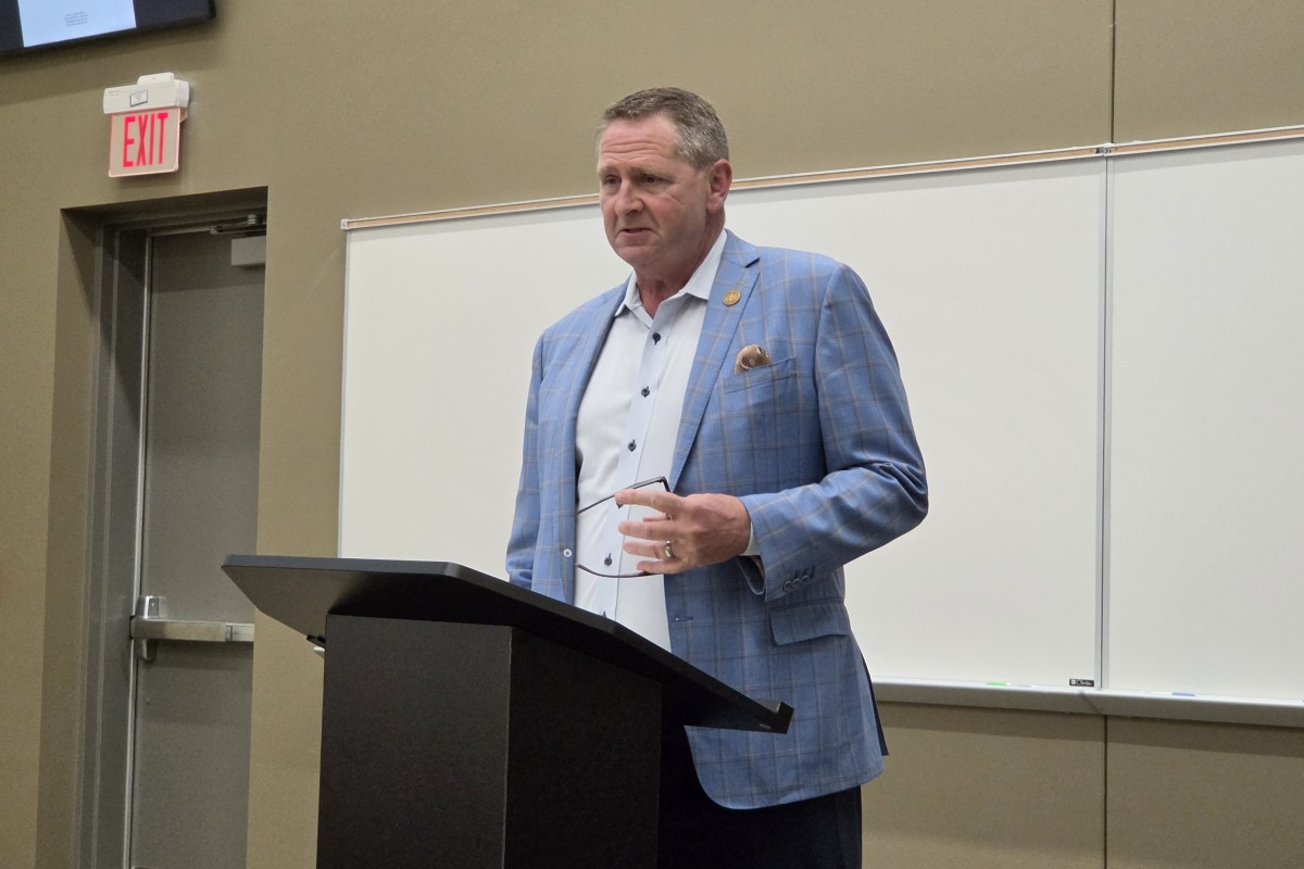 Man in a blue checkered blazer stands at a podium giving a lecture in a classroom setting.